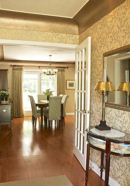 A well-lit dining room with wooden floors, green chairs around a table, large curtained windows, and a gold-accented entryway featuring shimmering effects from Modern Masters Metallic Paint on the mirror and table with lamps.