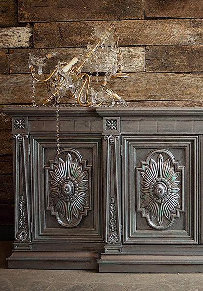 A dark wood cabinet with carved floral designs stands against a rustic plank wall. Atop it, a broken chandelier glimmers with shimmering colors achieved using Modern Masters - Metallic Paint - Gallon on its gold frame.