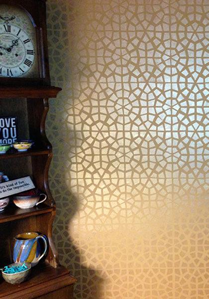 A wooden shelf with colorful pottery and a clock stands beside a wall painted with Modern Masters Metallic Paint, featuring an intricate geometric gold pattern that shimmers as light and shadows highlight the design.
