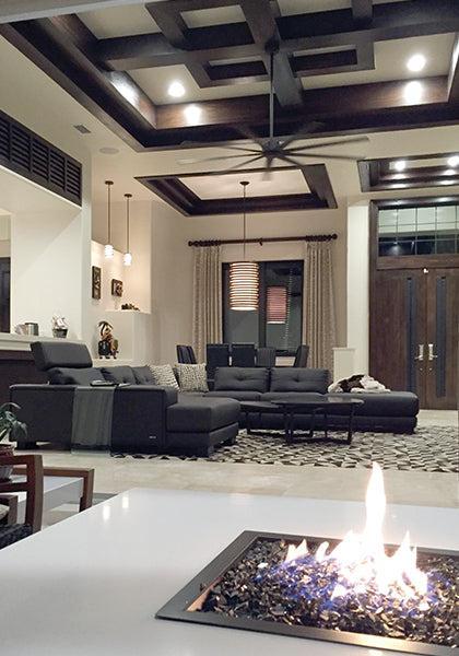 Modern living room with a geometric coffered ceiling, dark sectional, patterned rug, and glass fire feature. Warm lighting highlights wooden double doors finished in Modern Masters - Masterclear Supreme for lasting style.
