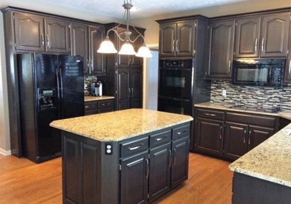 Modern kitchen features dark wood cabinets, black appliances, granite countertops, central island, and wood flooring protected with Modern Masters - Masterclear Supreme, plus pendant lighting and a mosaic tile backsplash.