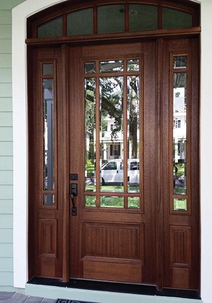 A wooden front door with glass panels and sidelights features decorative grids and is protected by Modern Masters - Masterclear Supreme. Reflections of trees and houses appear on the glass, framed by white trim around the porch.