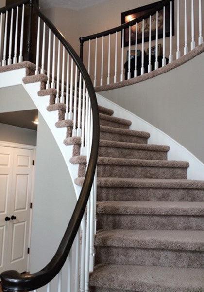 Curved staircase with beige carpet, white spindles, and a dark wood handrail finished in Modern Masters - Masterclear Supreme, leading to an upper floor with framed pictures and white double doors below.