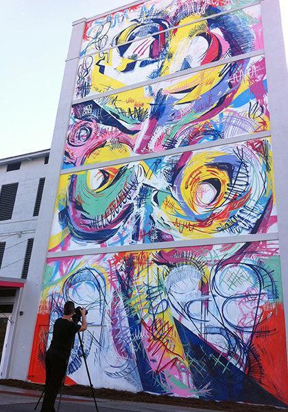 A person with a camera tripod photographs a tall, colorful mural of abstract swirls and vibrant spots on a building wall, protected by Modern Masters - Masterclear Supreme, a durable aliphatic polyurethane topcoat.