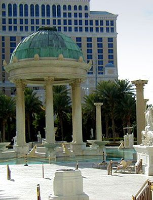 A domed pavilion with columns coated in Modern Masters - Metal Effects Patina Aging Solution, showing bronze hues and blue-green patina, stands by a pool with statues, palm trees, and a multi-story hotel in the background.