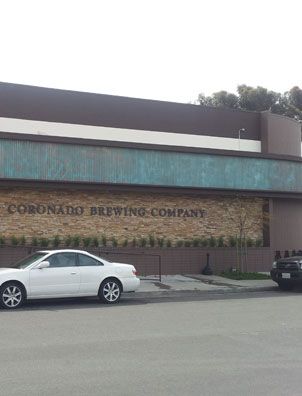 A white car is parked by a building with a sign reading “Coronado Brewing Company” on a stone wall showing green or blue hues, created using Modern Masters Metal Effects Patina Aging Solution. Shrubs line the base and the sky looks cloudy.