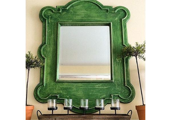 A wall displays a green-framed mirror with a faux finish created using Modern Masters - Tintable Glaze, flanked by potted plants. Beneath, a wooden shelf holds four glass candle holders in a row.
