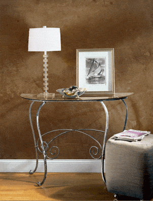 A small glass-top table with decorative metal legs holds a modern white lamp, sailboat photo, and silver bowl. Nearby is a gray ottoman with magazines atop, set against a brown wall finished with Modern Masters - Venetian Plaster Topcoat.