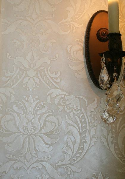 A close-up of durable, cream-colored Modern Masters ShimmerStone wallpaper with a damask pattern and decorative finishes, paired with a wall sconce featuring a lit candle and crystal ornaments on the right.