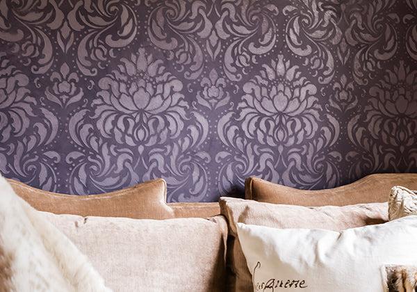 A beige sofa with plush cushions sits before a wall finished in Modern Masters - ShimmerStone, showcasing ornate purple damask patterns and decorative finishes for an elegant touch.