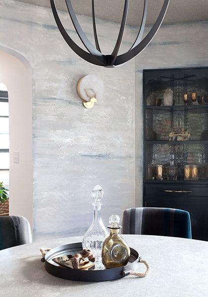 A round table with a tray of decanters and snacks sits under a modern black chandelier. The textured wall features durable Modern Masters - ShimmerStone decorative finishes, with a sconce and dark cabinet with glassware in the background.