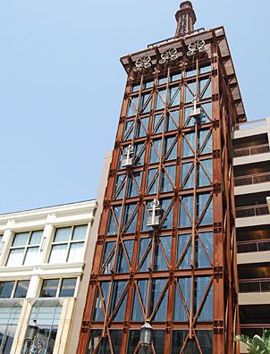 A tall clock tower with exposed steel framework and large mechanical clocks features a weathered finish created using Modern Masters - Metal Effects - Rust Activator, standing beside a white building under a clear blue sky.