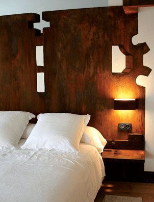 A bed with a white comforter and pillows stands against a dark wooden headboard featuring interlocking puzzle pieces finished with Modern Masters - Metal Effects - Rust Activator. A warm lamp is mounted above a small shelf on the right side.