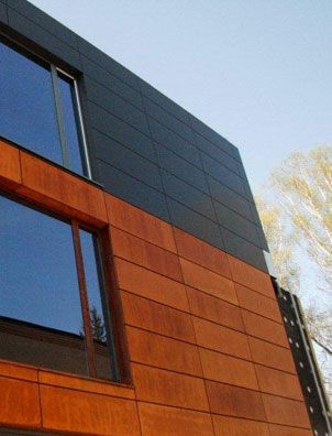 A modern facade features sleek rectangular panels; the lower section uses Modern Masters - Metal Effects - Rust Activator for a weathered rust look with wood-like accents, while the upper part has dark smooth panels. Large windows reflect sky and trees.